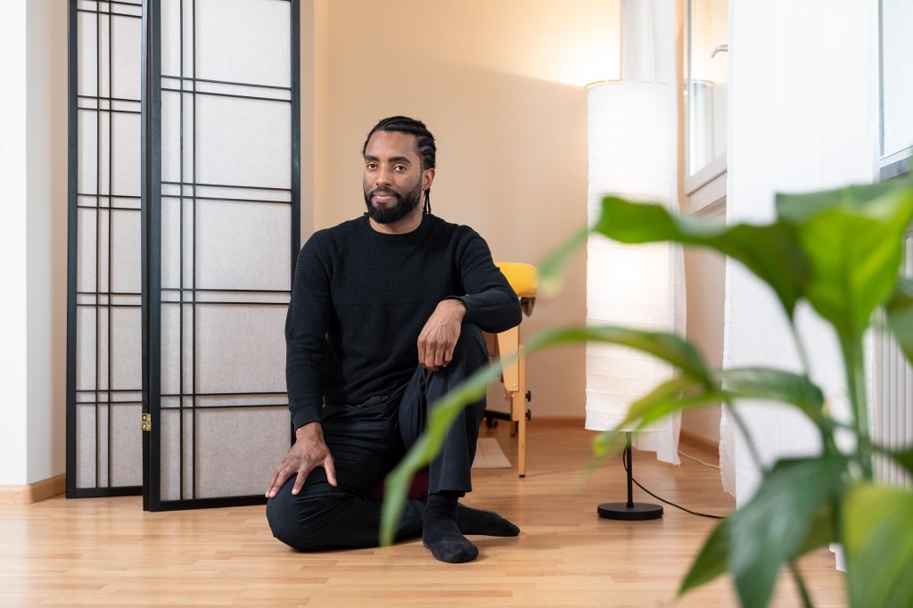 Porträt von Daniel Fernandes.
Daniel sitzt im Schneidersitz auf einem Yoga Kissen. Links von ihm befindet sich eine Zimmerpflanze mit grossen Blättern.